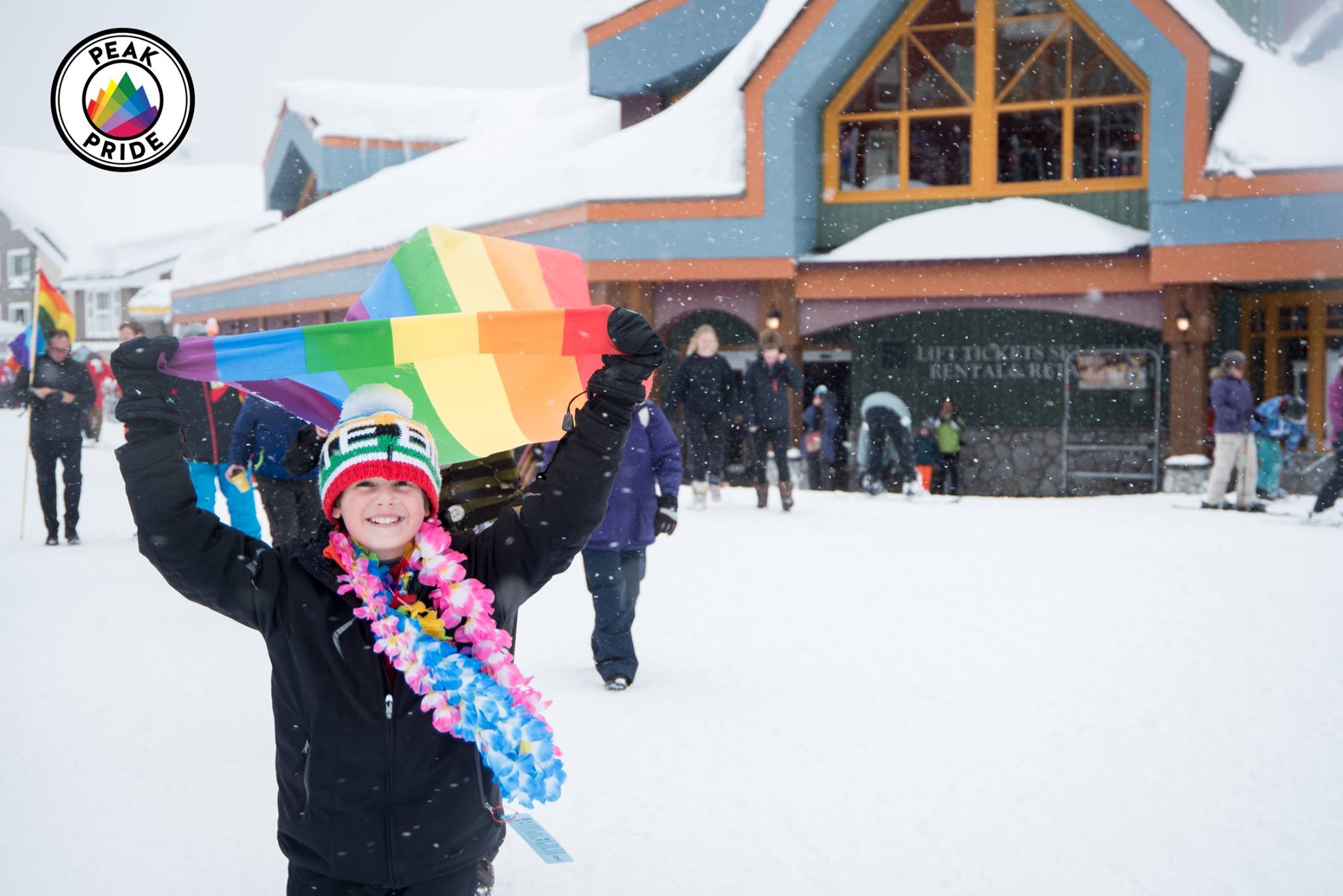A smiling youth holds a pride flag while wearing winter clothes. Snow is falling.