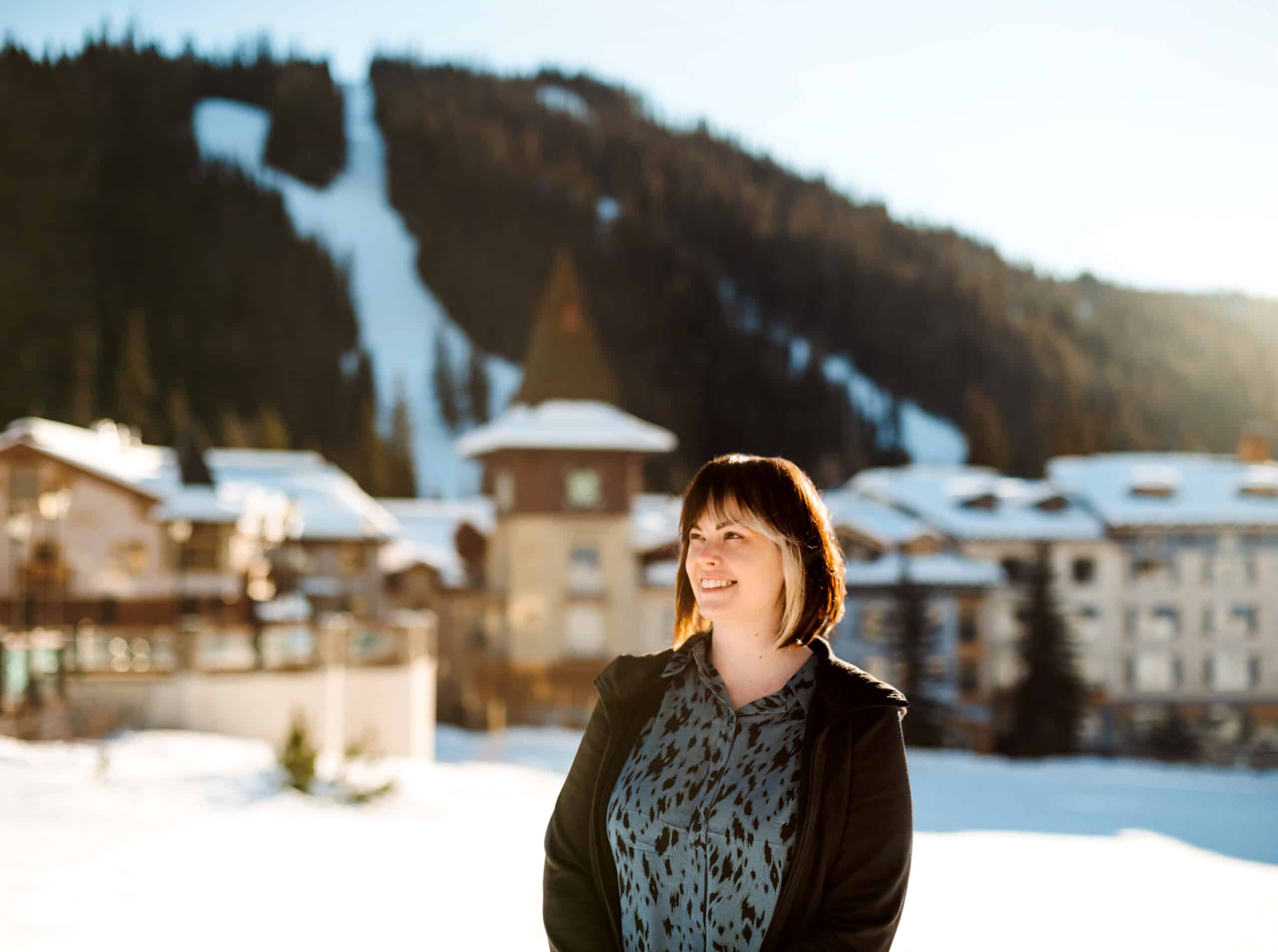 Brandi, a white woman with brown hair, stands outside in Sun Peaks. Village buildings and the ski runs are visible behind her.