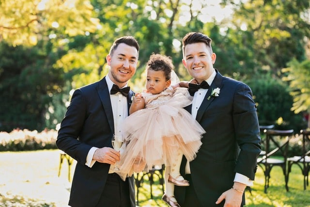 A couple from Ireland named Mark and Damien stand outside holding their daughter, Izzy, while wearing suits and smiling. 