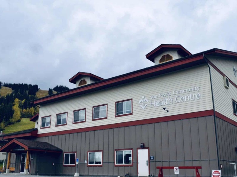 Sun Peaks Community Health Centre is a large beige, grey and red building. A grey sky, green ski slopes and trees are seen in the background.