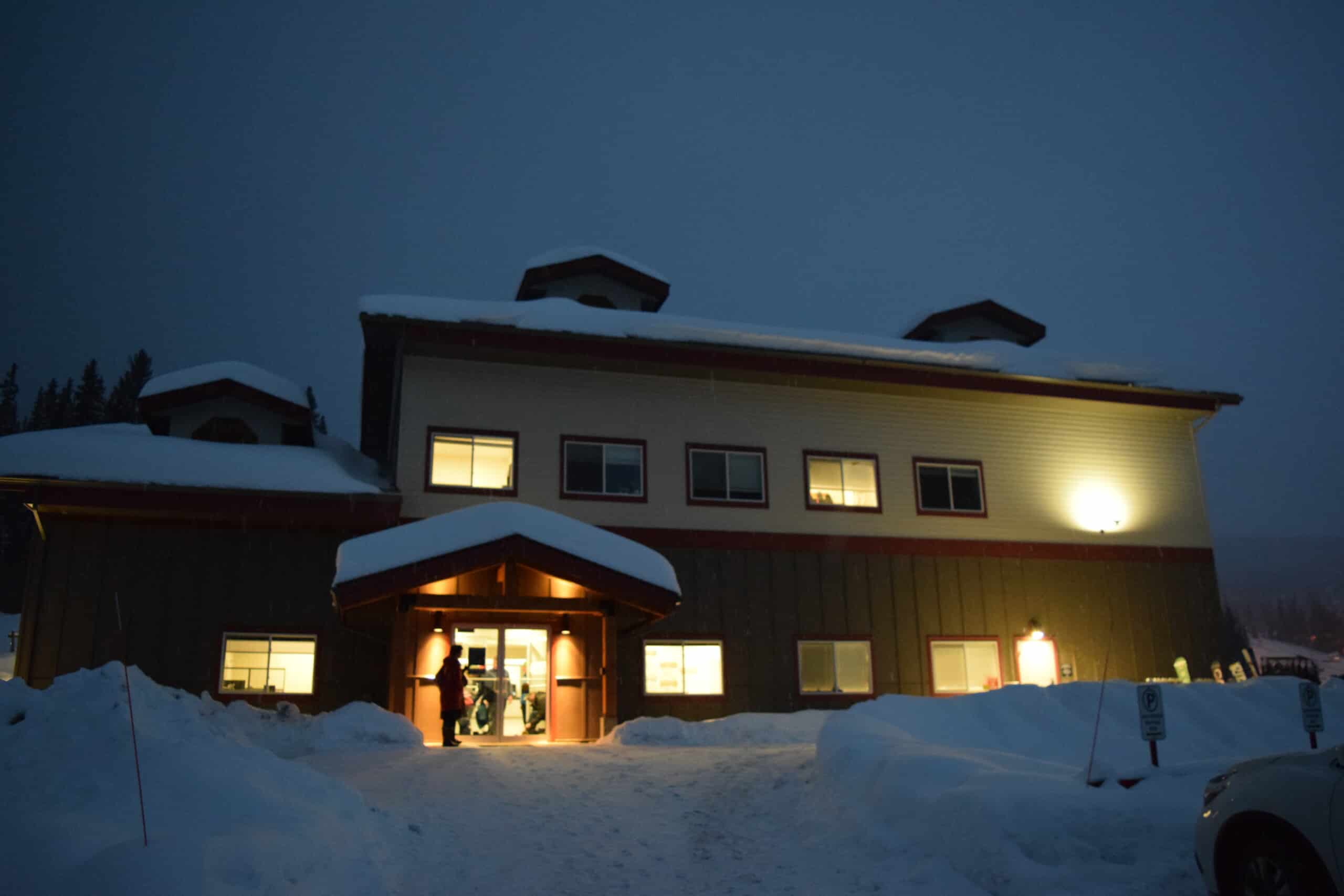 Sun Peaks Community Health Center is a beige building with snow in front of the entrance. Warm yellow light glows from the windows.