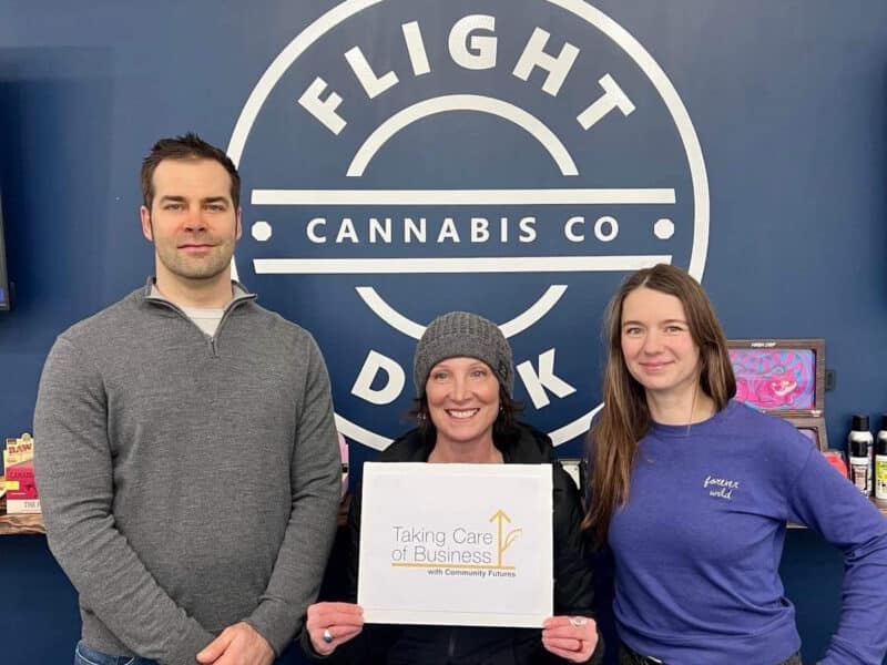 Kyle Vike, Julie Kimmel and Sonya Trevisi smile and stand in front of a blue wall with a white logo that reads "Flight Dek Cannabis Co." Kimmel is holding a white sign that says "Taking Care of Business with community Futures."