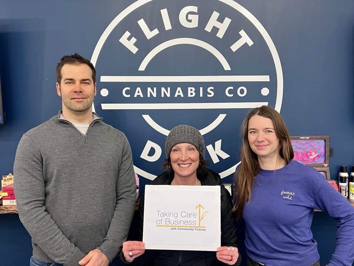 Kyle Vike, Julie Kimmel and Sonya Trevisi smile and stand in front of a blue wall with a white logo that reads "Flight Dek Cannabis Co." Kimmel is holding a white sign that says "Taking Care of Business with community Futures."