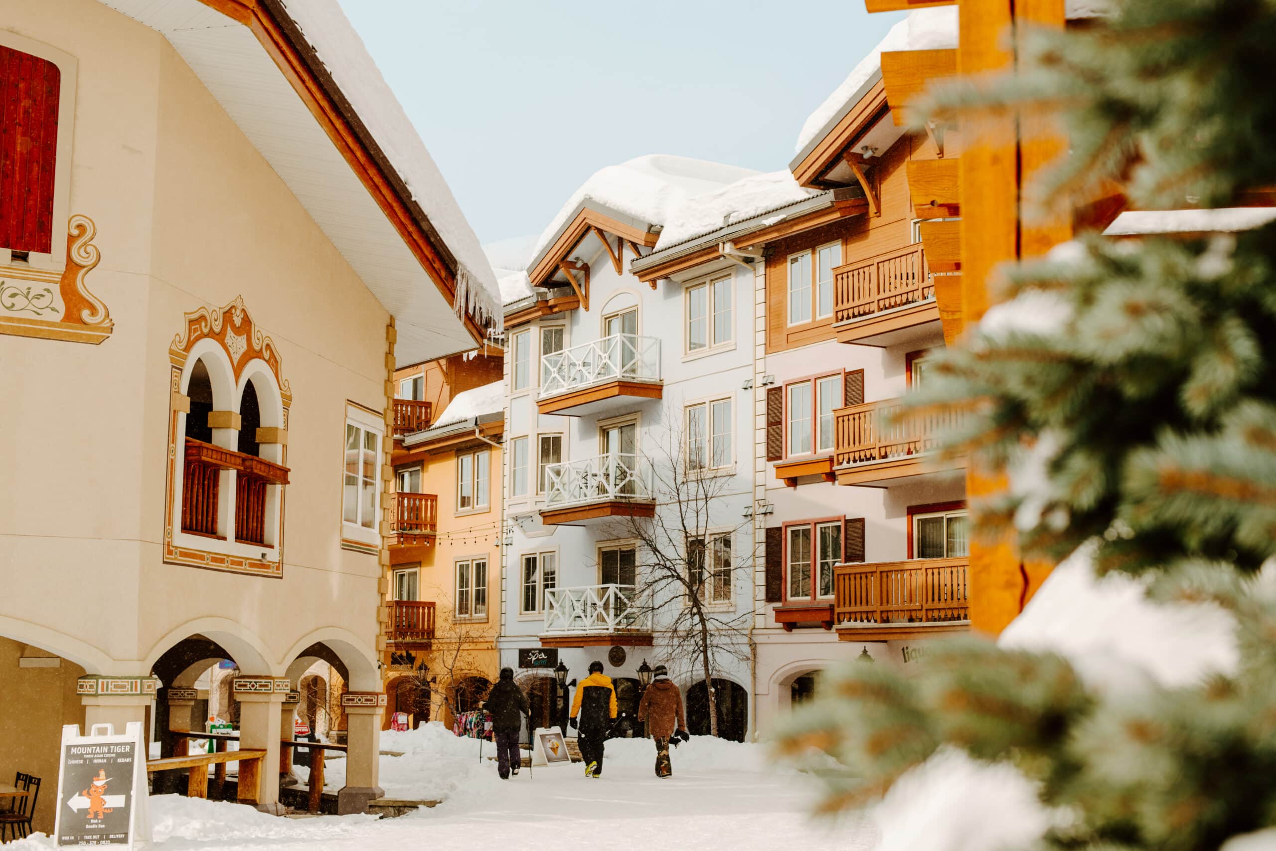 Three storey high Swiss Alps style buildings surround a walkway covered with snow.  Three people wearing ski and snowboard clothing are in the centre walking with their backs visible.