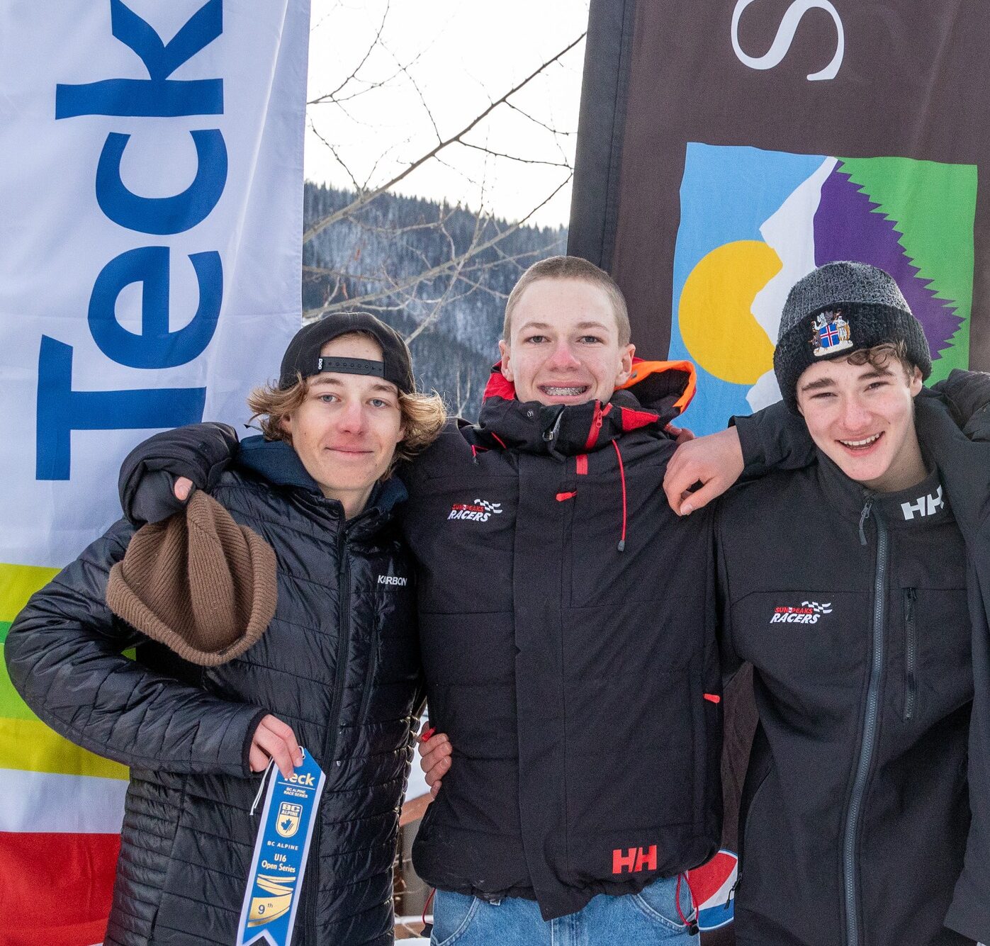 Three teen boys, members of the Sun Peaks Alpine Club, stand together in front of event banners after the U16 Teck Open event.