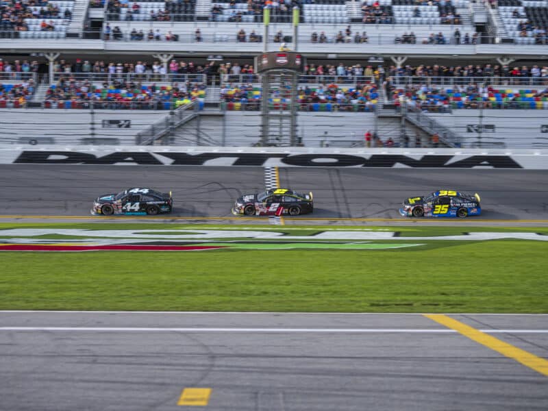 Three racecars speed along a racetrack in Daytona Beach, Florida. Jason White is in car #44,leading the race. Fans are in the stands in the background of the image.