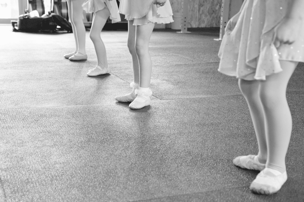 A black and white photo of four children in ballet skirts in the Sun Peaks Academy of Dance studio. Only the children's legs are shown.