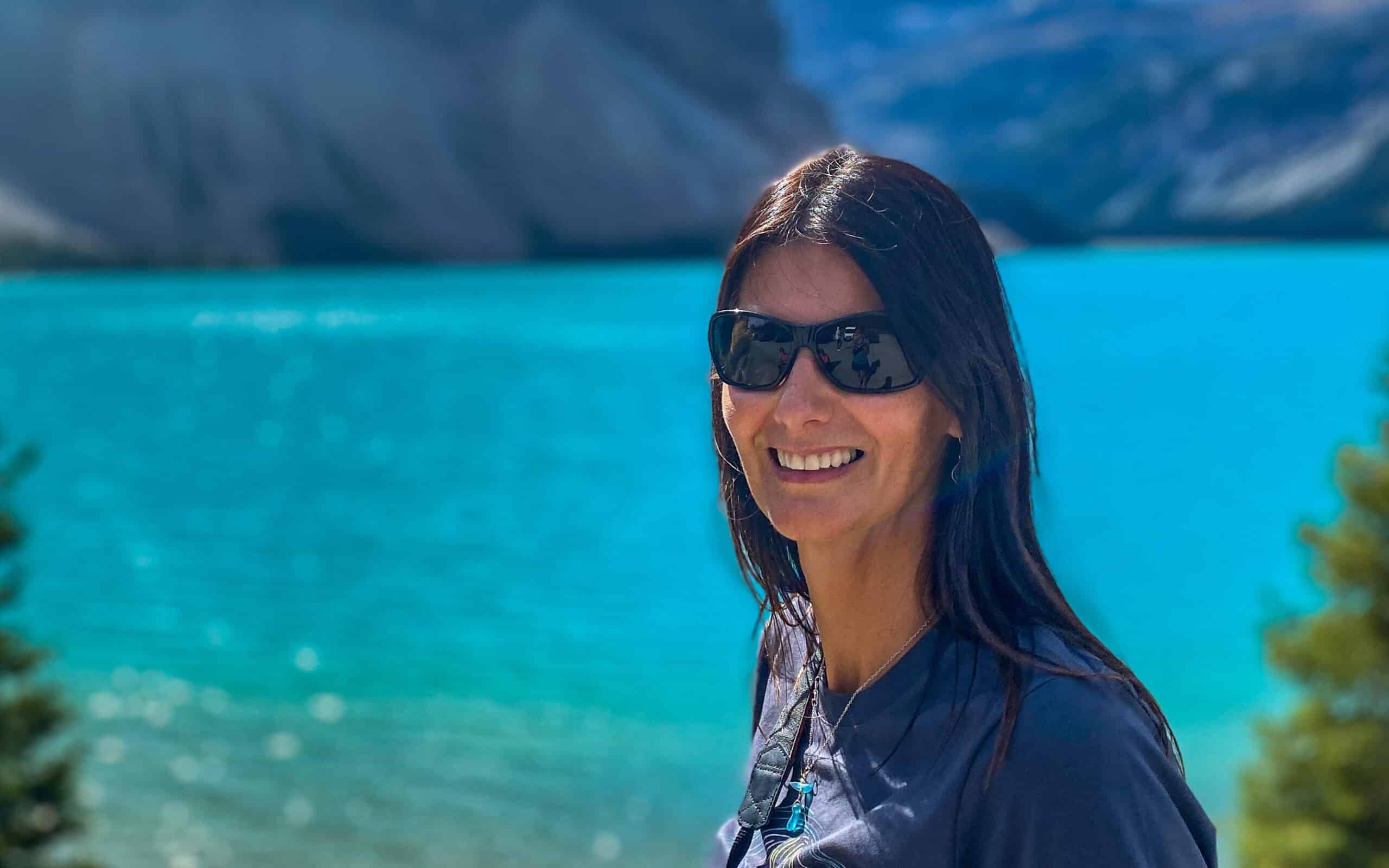 Maria Davis hasmedium length brown hair and is wearing sunglasses. She's smiling at a camera. The background features a bright blue lake with mountains in the background