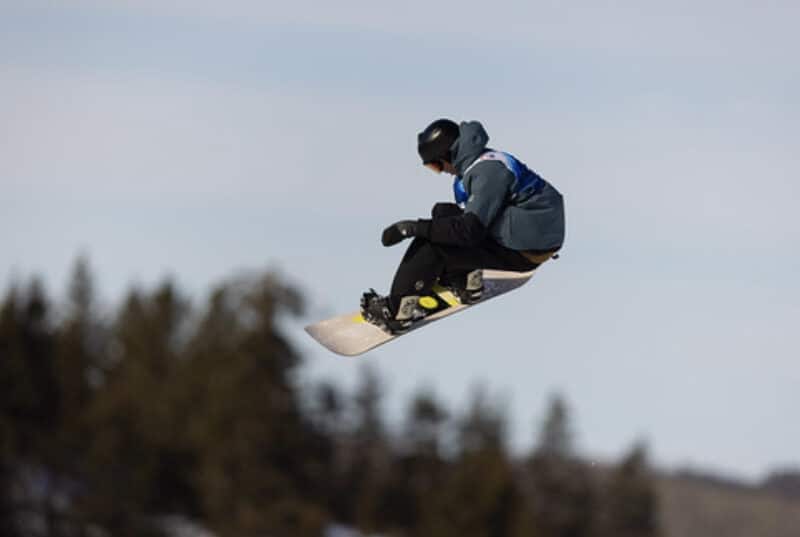 Snowboarder Nayte Brown catches air in P.E.I. at the Canada Winter Games. A grey sky and trees are behind him. He's wearing a teal jacket and black pants with a white and yellow snowboard.
