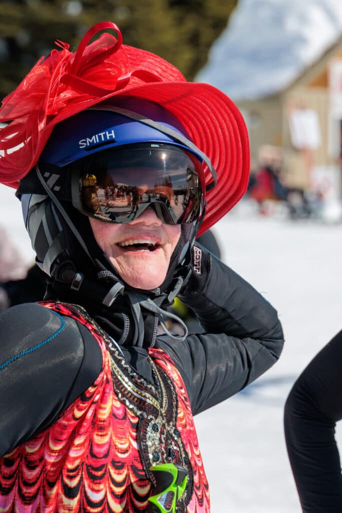 A woman wearing a red straw hat over her helmet and a red bedazzled dress poses for the camera and laughs.