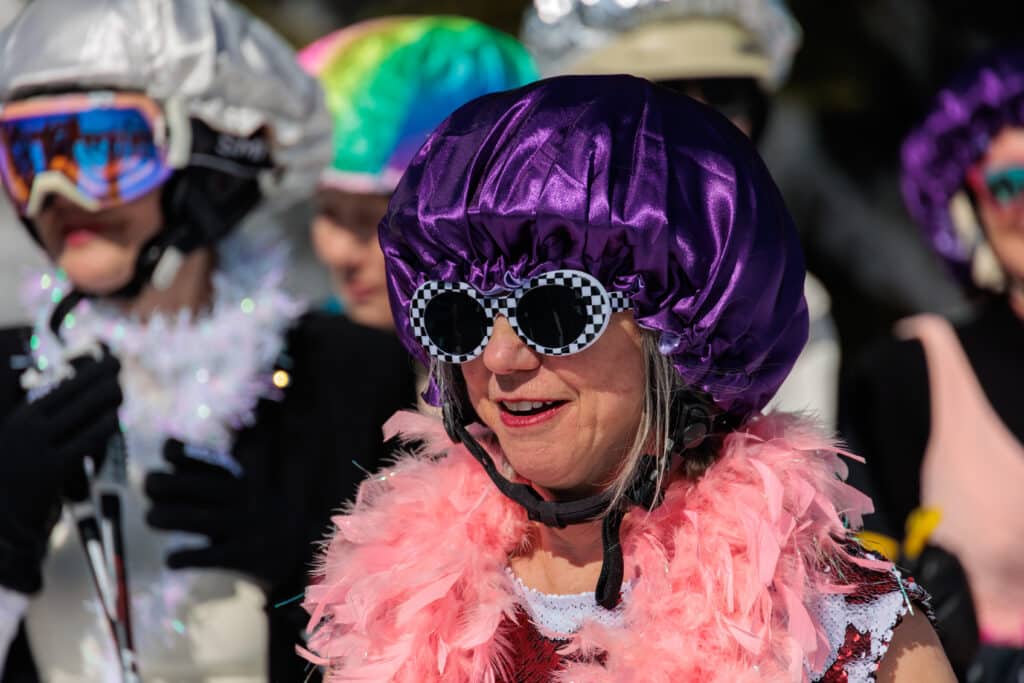 A woman wearing a purple bonnet, checkered sunglasses and a pink feather boa smiles.