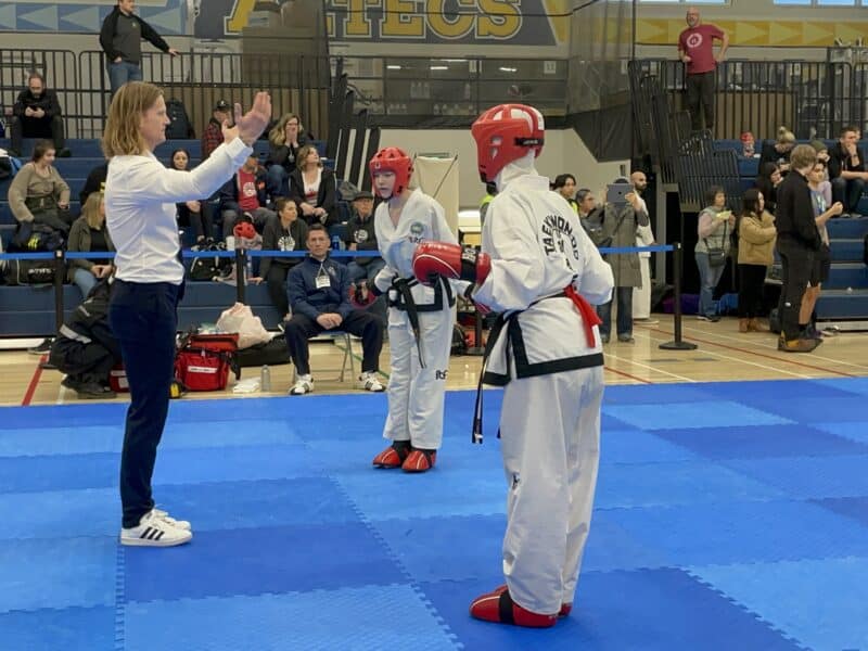Taekwon-Do athletes stand on a blue mat in a gymnasium at Alpha Secondary School in Burnaby. They are competing in provincial championships and wearing white dobok uniforms with red gloves, helmets and shoes. Coach Ryan Oevermann sits in a chair on the sidelines wearing a blue sweater, black pants and white shoes with three black stripes. There is a small crowd of people in the stands.