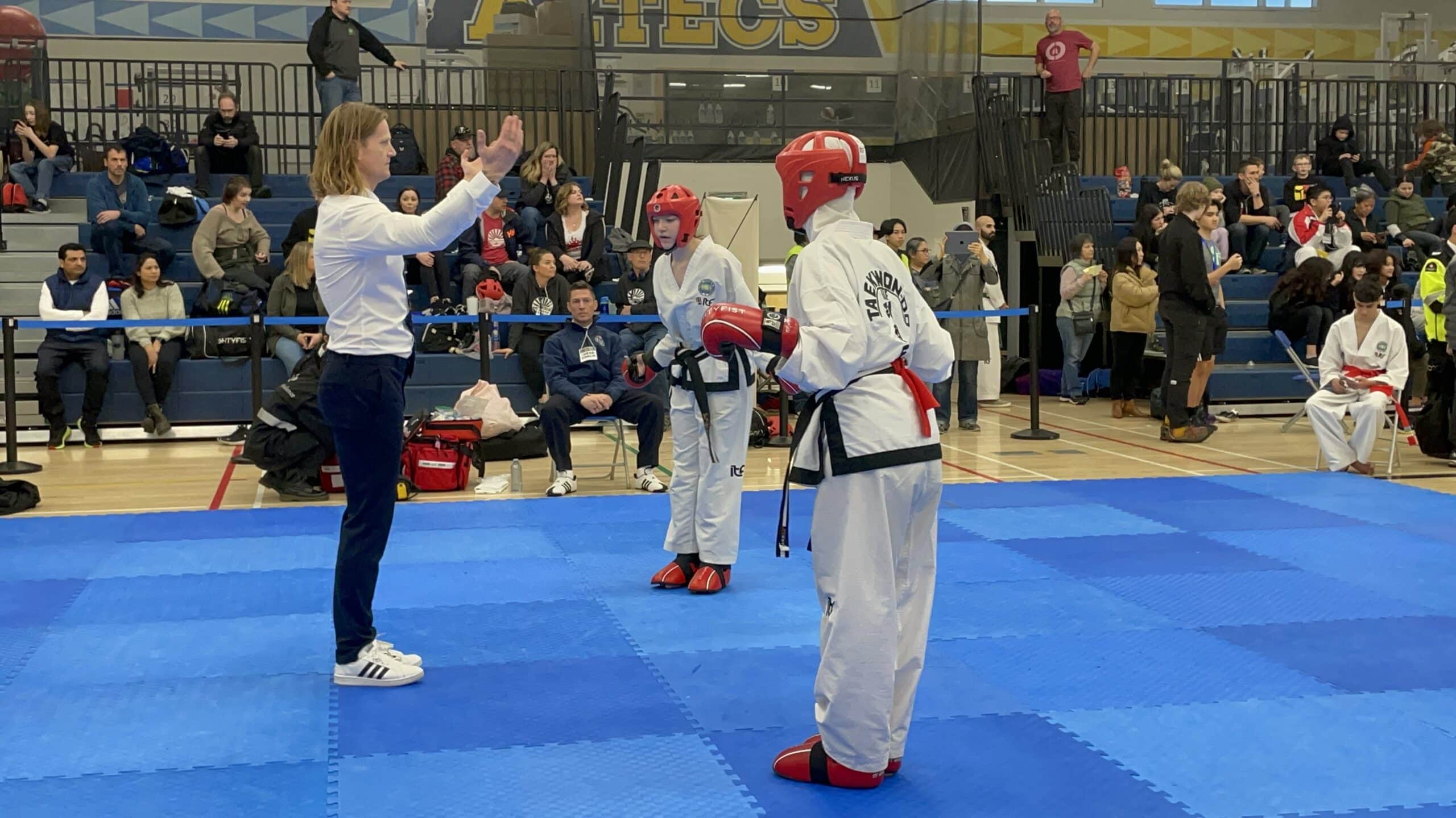 Taekwon-Do athletes stand on a blue mat in a gymnasium at Alpha Secondary School in Burnaby. They are competing in provincial championships and wearing white dobok uniforms with red gloves, helmets and shoes. Coach Ryan Oevermann sits in a chair on the sidelines wearing a blue sweater, black pants and white shoes with three black stripes. There is a small crowd of people in the stands.