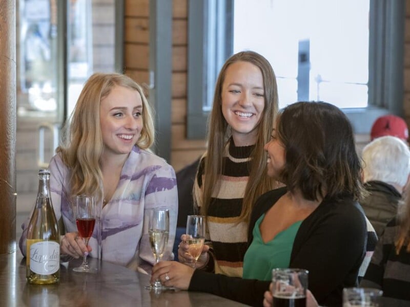 Three smiling women look at each other while holding glasses of wine at a table.