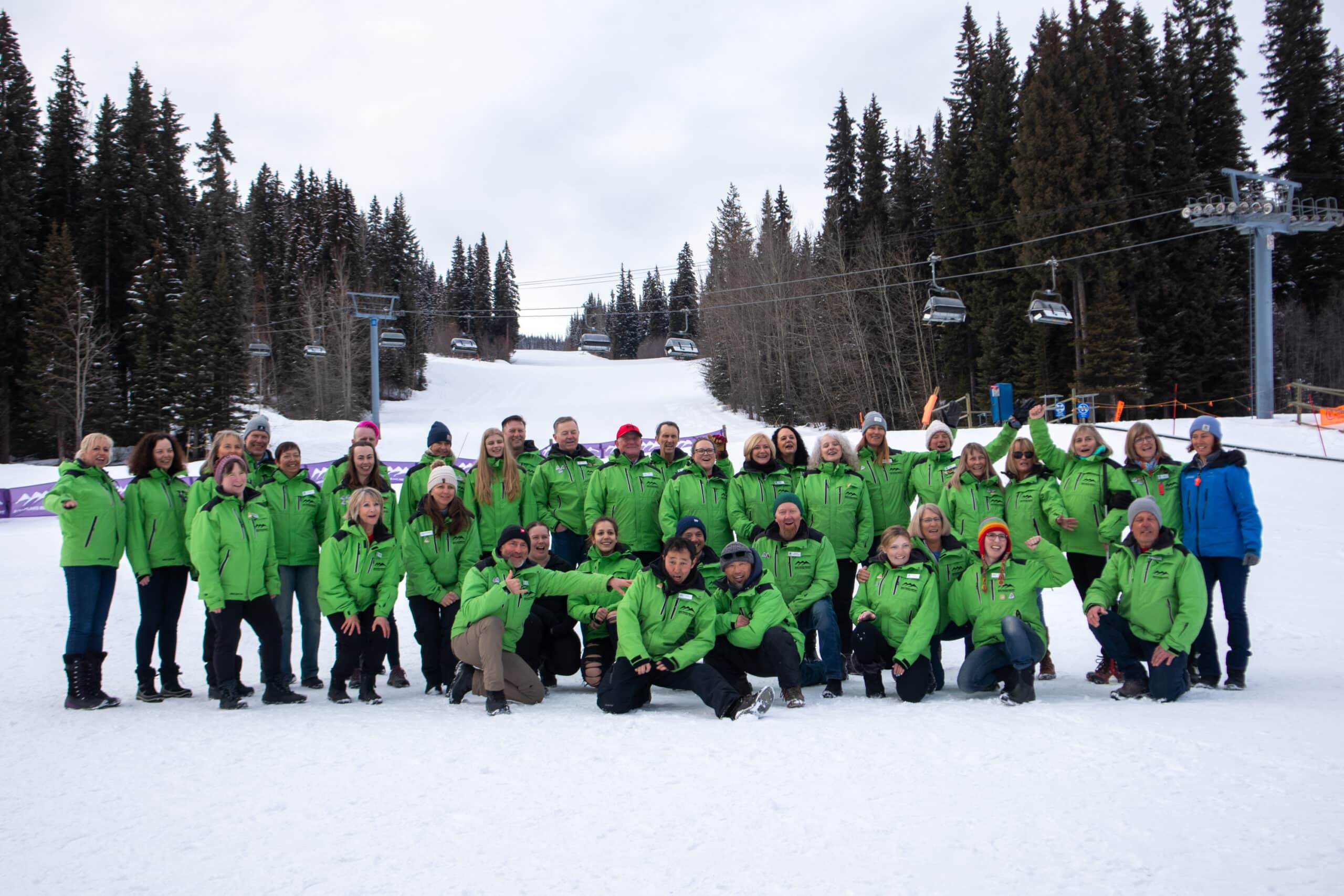 A large group of ski instructors wearing green jackets gather for a photo in front of a ski hill. There is a chairlift in the background.
