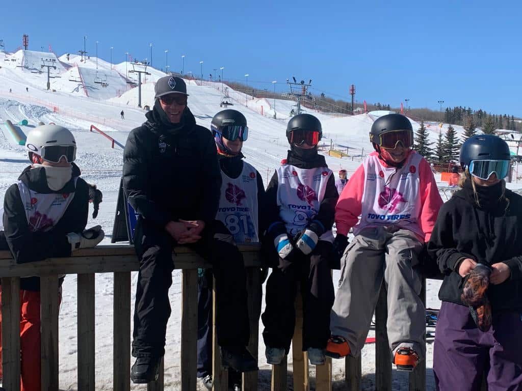 Five competitive skiers and one Sun Peaks Freestyle Club coach stand together outside with blue skies and a ski hill in the background.