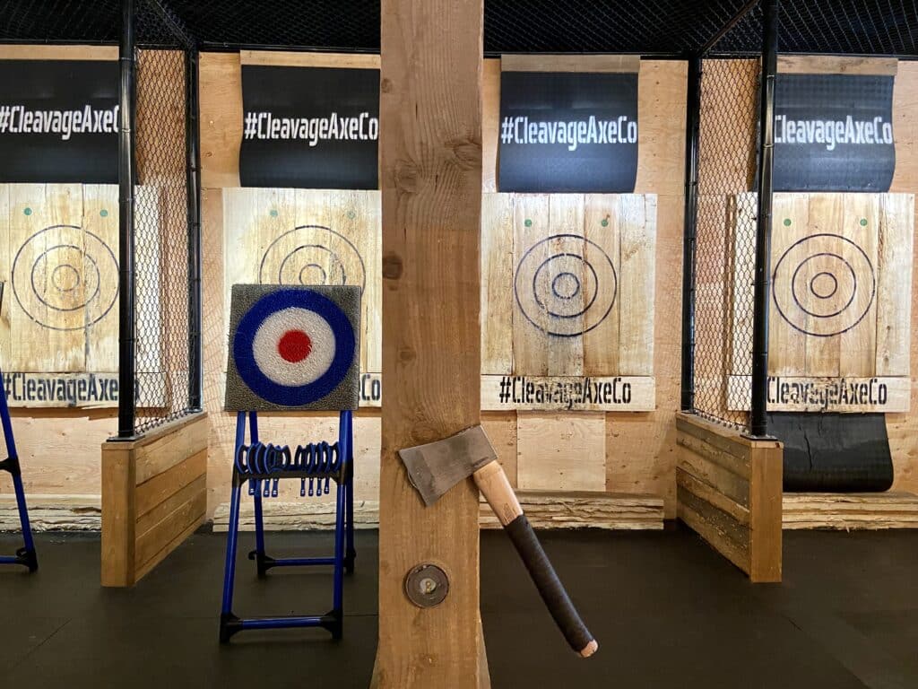 Four wooden bulls-eye signs with indents from axes are in a line. Fencing seperates the targets. Black signs with a hashtag and the words "Cleavage Axe Co" are at the top of the signs. An axe is in the centre of the photo on a wooden pillar.