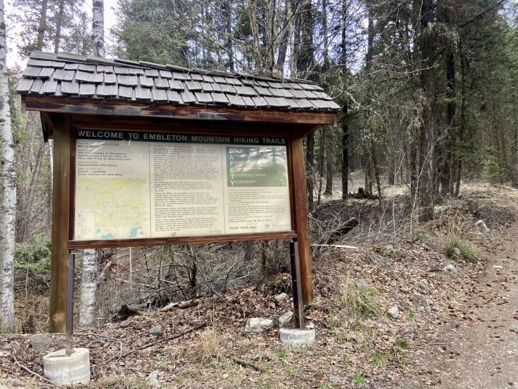 A large sign is in the left hand side of this picture. The words "Welcome to Embleton Mountain Hiking Trails" are at the top of the sign. There is a map and description of the area. Leaves and grass dot the ground, with trees behind the sign. A dirt road is in the right hand lower corner of the image.