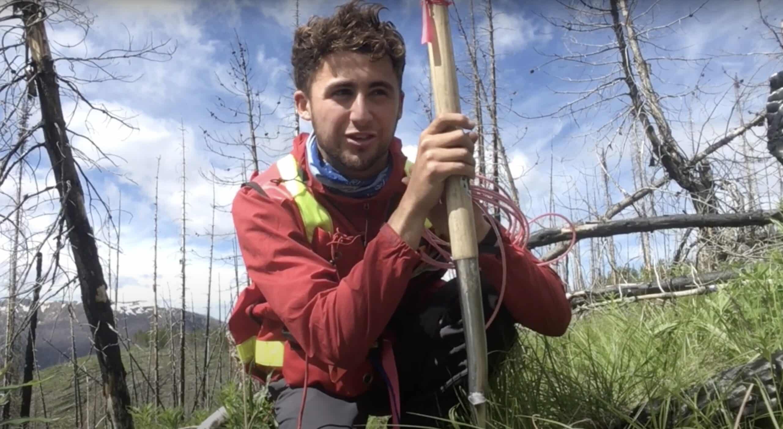 A man wearing a red and yellow jacket holds a tool for digging while grouching in grass. He is researching Sun Peaks bears through their feces. A blue sky and dead trees are in the background.