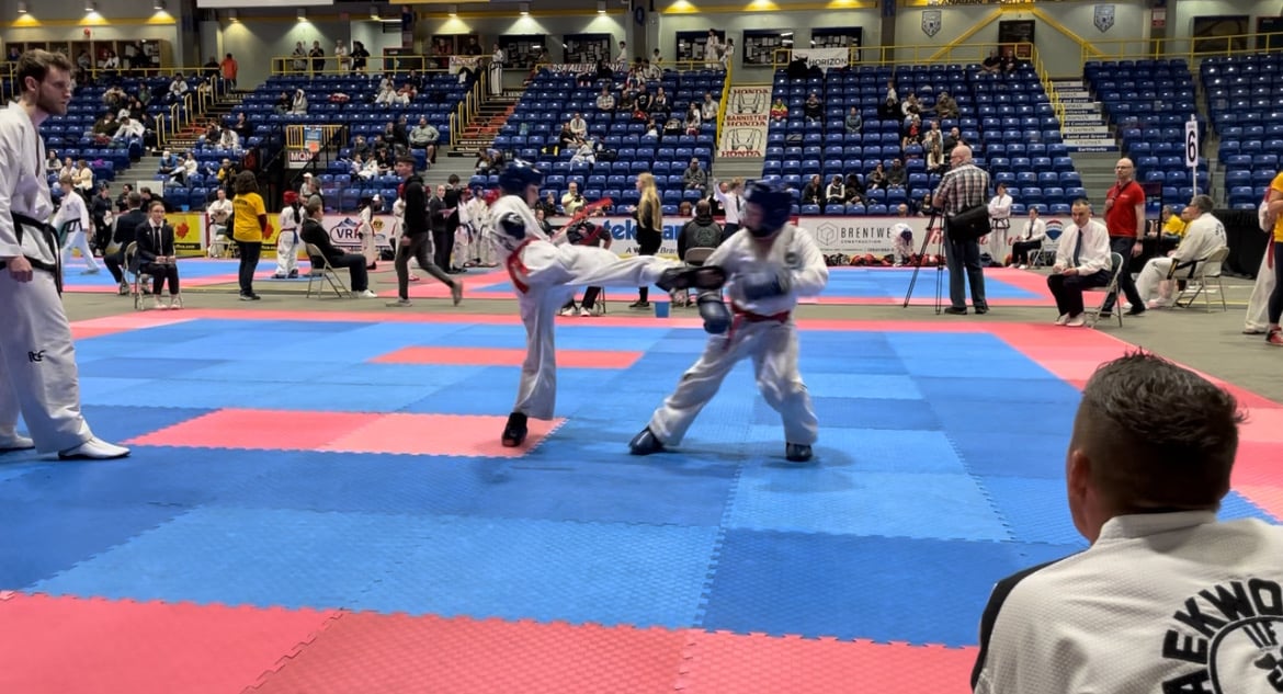 Two taekwon-dp athletes fight on a red and blue mat inside a large gym. A coach and a referee watch them. There are blue seats in the stands located in the background of the photo with audience members in some of the chairs.