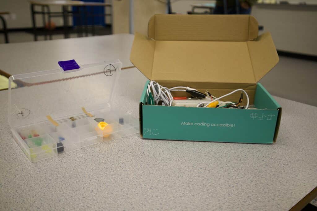 A blue cardboard box holding items used for making microbits is in the centre of the photo. A plastic case holding more items beside it. The boxes sit on a table inside a school portable.