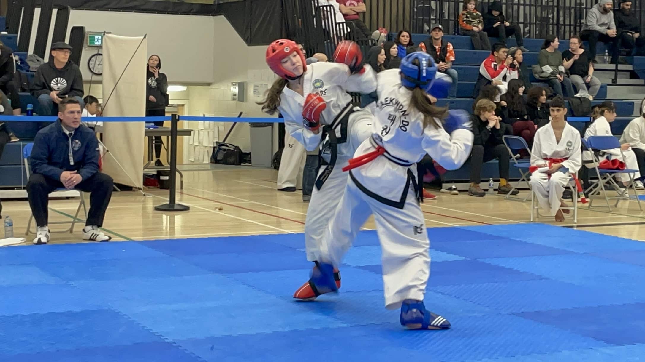 Two women including Montana Oevermann are wearing white robes are in the centre of the photo. They are competing against each other in a taekwon-do competition. One has a red helmet, gloves and shoes. The other has a blue helmet, glove and shoes. They are on a blue mat. There is an audience in the stands in the background.