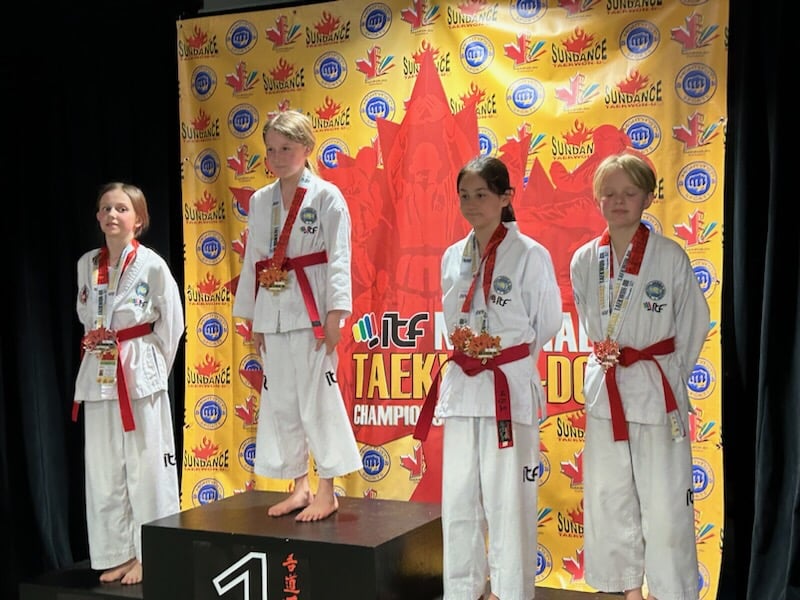 Four youth from the Northern Taekwon-Do Academy stand on podiums while wearing white uniforms called doboks. They stand in front of a yellow square banner for a tekwon-do competition. They each have medals hanging from their necks.