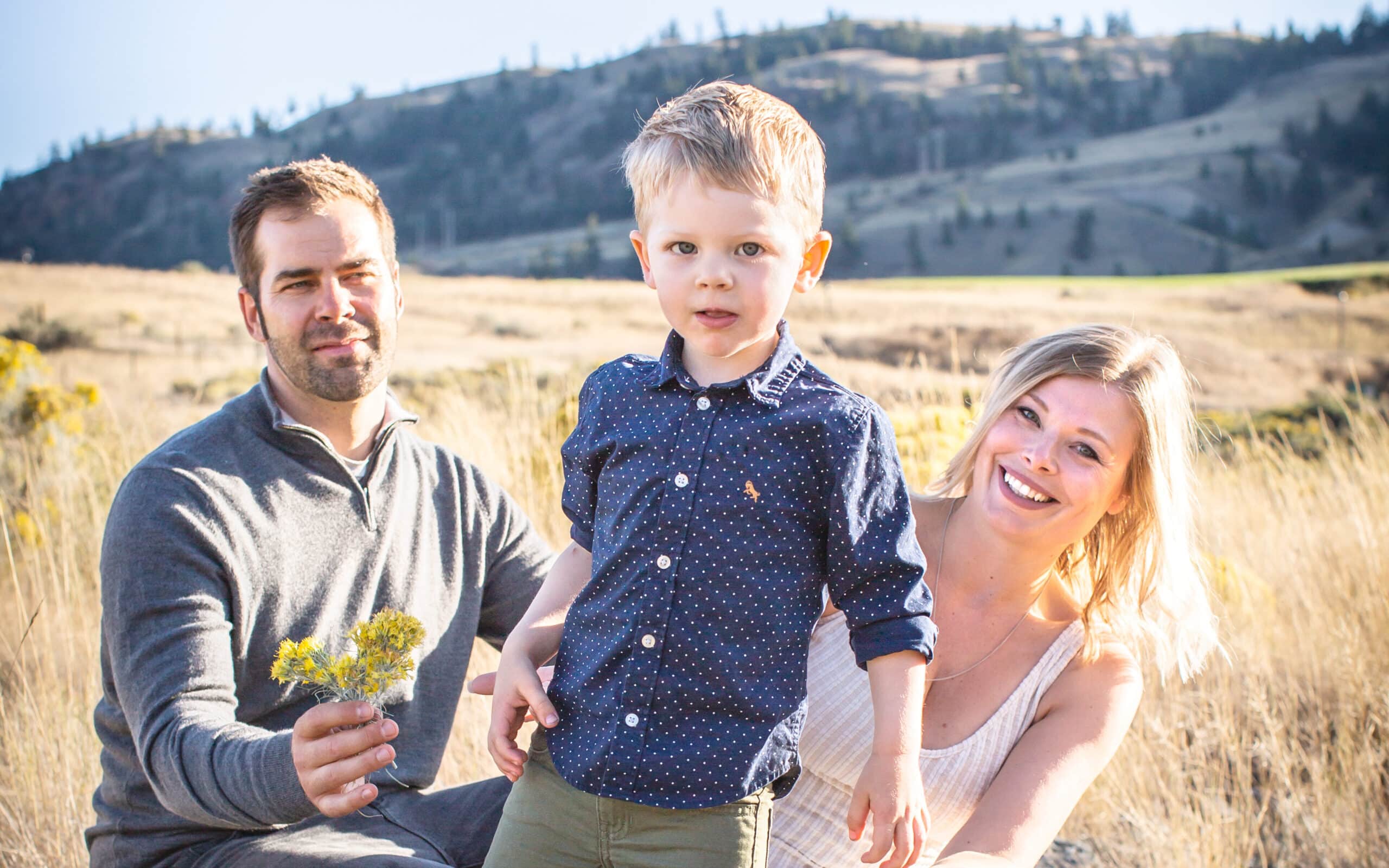 A child with blonde hair and blue eyes is in the centre of the photo. He's wearing a blue t-shirt with polka dots. His parents kneel behind him. The dad has short brown hair and wears a grey sweater. His mom wears a white tank top and has medium length blonde hair. Grasslands and rolling hills are in the background.