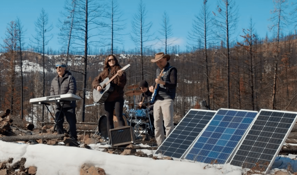 Three people play instruments on a stage outside. There are mountains in the background and snow on the ground. It is a sunny day.