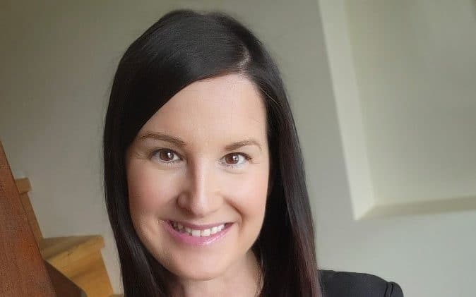 A woman with light skin and brown eyes smiles at a camera. She has straight brown hair and there's a white wall behind her. She will be the New chief administrative officer.