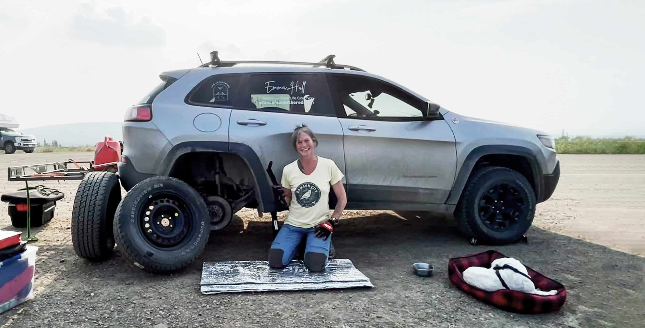 A blonde woman squats in front of a silver SUV. She's holding a wrench to change a tire. A dog sits in a red bed beside her. There is a dirt ground in the foreground and a smoky sky in the background.