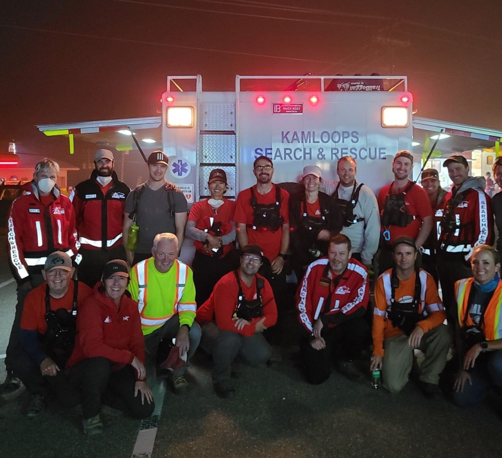 This photo was taken at night, Kamloops Search and Rescue (KSAR) volunteers pose together in front of an ambulance with its lights on.