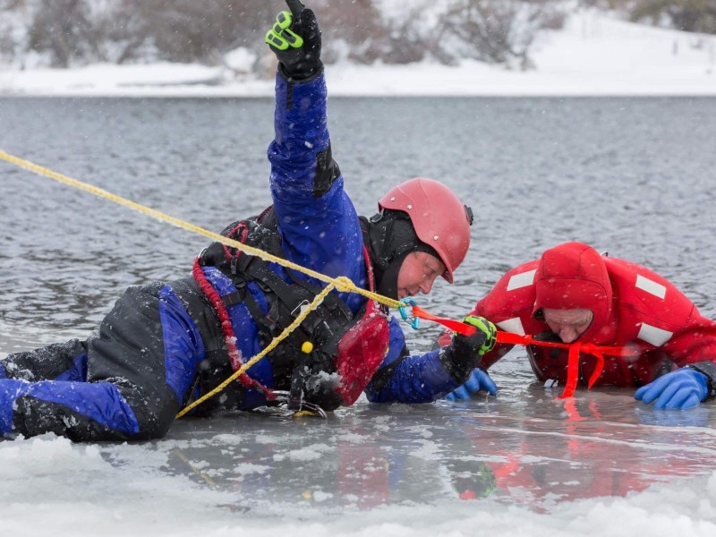 A Kamloops Search and Rescue (KSAR) volunteer lays on ice rescuing someone in red.