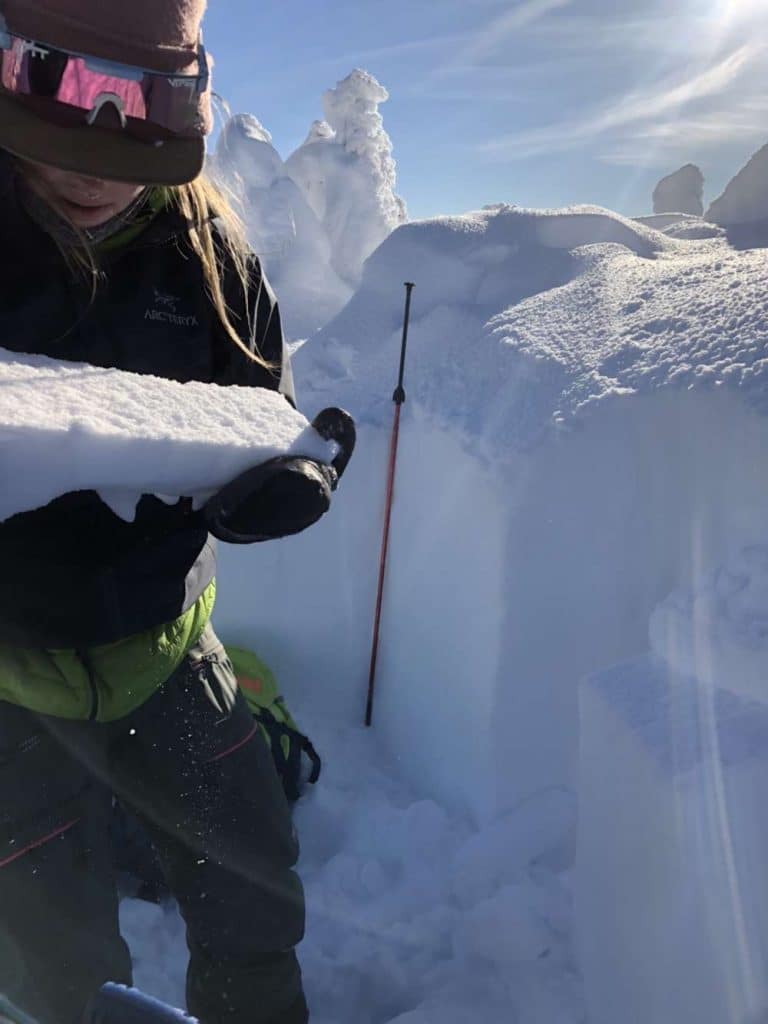 A woman inspects a rectangle of snow to determine avalanche risk. She's surrounded by deep snow and there's a blue sky in the background.