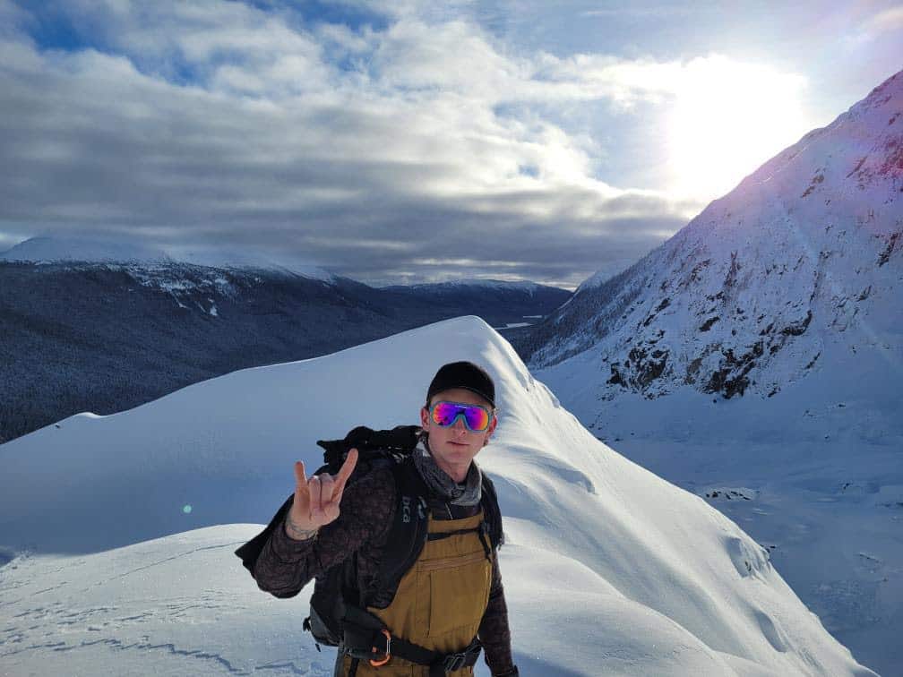 A man wearing sunglasses stands in a mountainous and snowy landscape. The sun shines in the background.