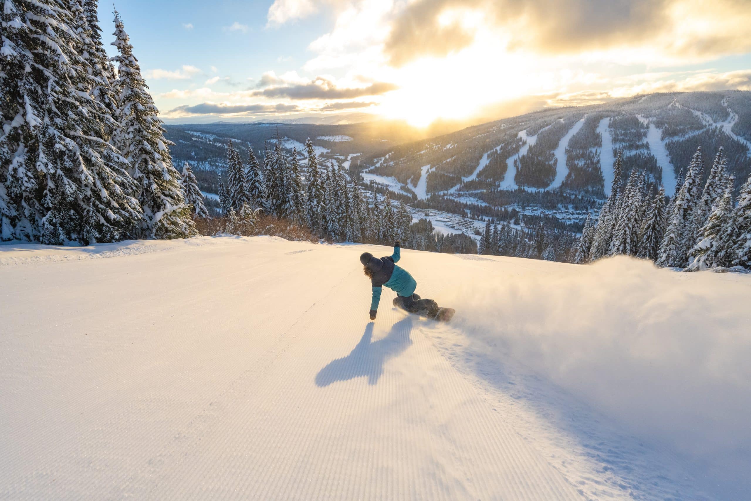A snowboarder is in the centre of the image and rides down a groomed ski run. A trail of powder flies up from the rider on the right side of the picture. The background features a sunset over a mountain with ski runs in the distance.