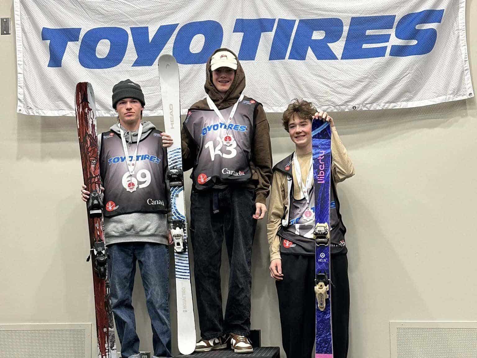 Three teenagers stand on a podium holding skis with a sign that says "Toyo Tires" in the background.